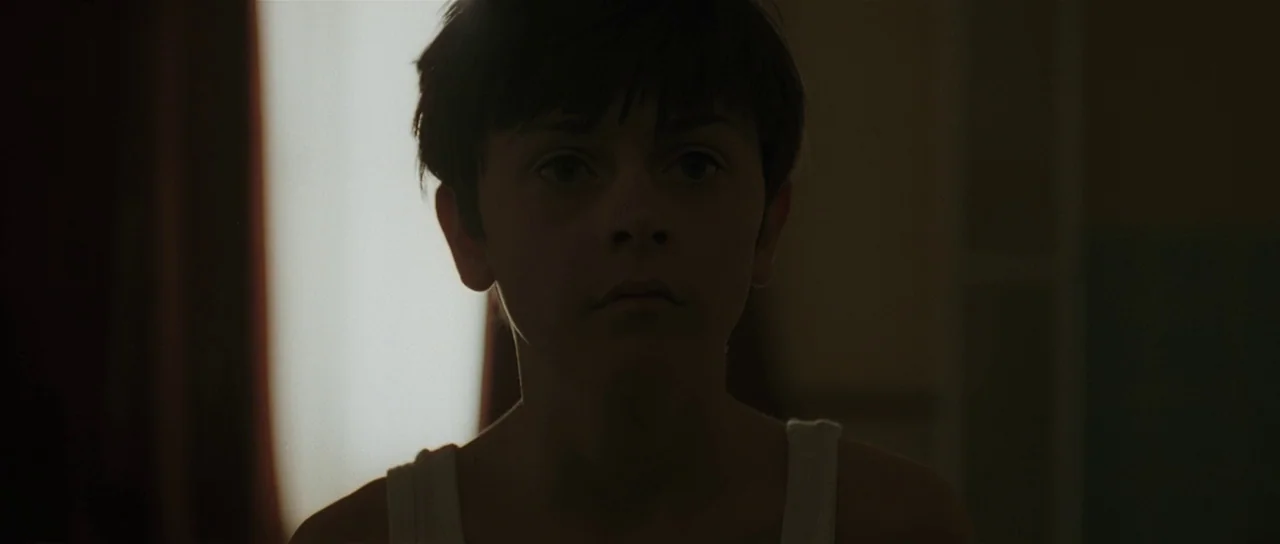 Bullhead (2011) movie still: closeup — A dimly lit room with a bright window visible behind the subject. A young boy in a white tank top stands…