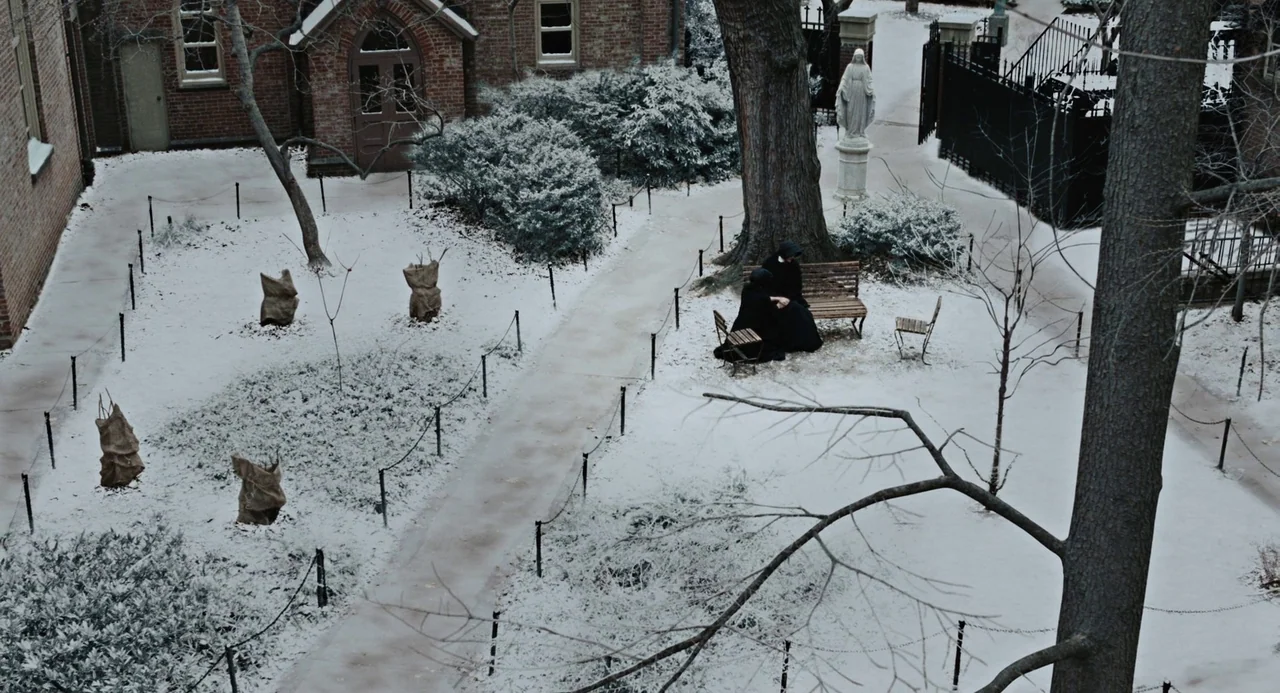 Doubt (2008) movie still: wide — A snow-covered cloister courtyard features a brick building and a central tree-lined walkway. Two figures dressed…