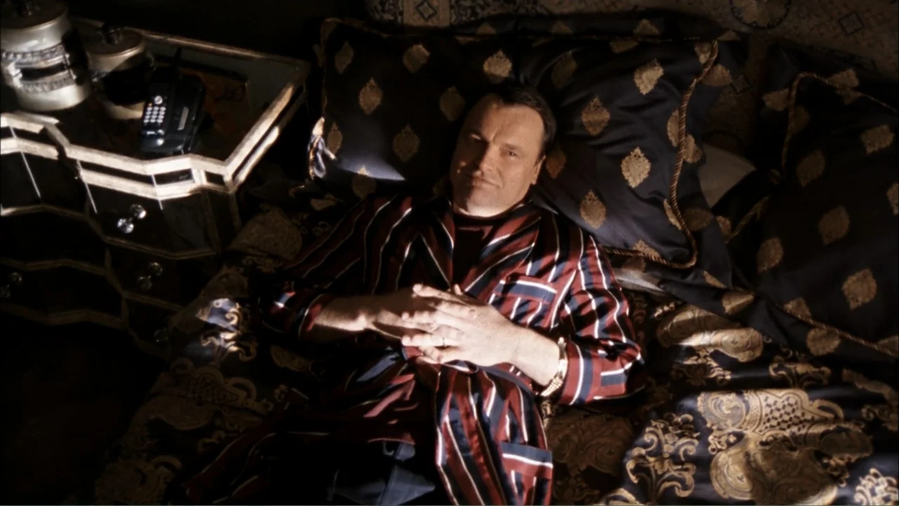 Driven to Kill (2009) movie still: medium — A dark bedroom with ornate dark patterned bedding and a mirrored side table. A man in a striped silk…