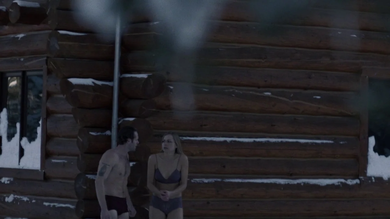 Snowbound (2017) movie still: medium — A rustic log cabin with snow-covered logs stands in an outdoor winter environment. Two people in minimal…