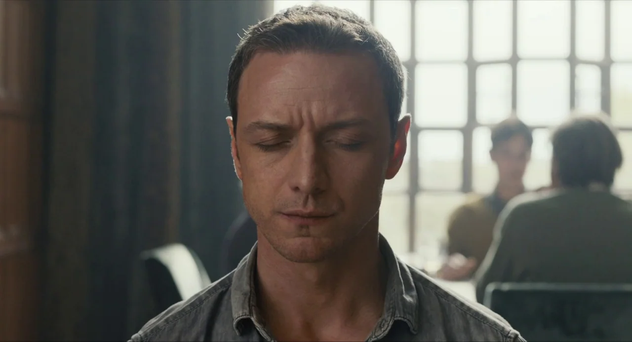 Submergence movie still: closeup — An interior space with a large grid-pattern window and dark wooden wall paneling. A man in a grey shirt sits in…