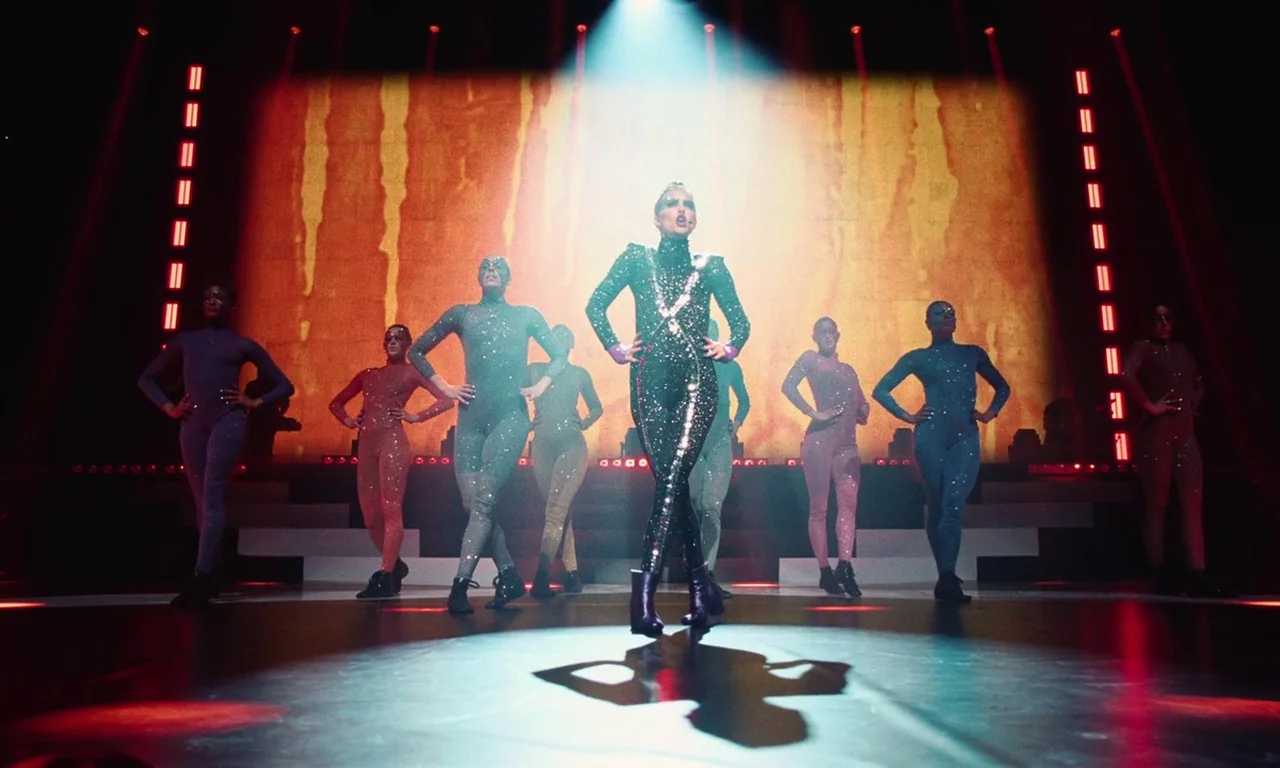 Vox Lux (2018) movie still: fullbody — A brightly lit stage features a performer center under a singular intense white spotlight. Several dancers…
