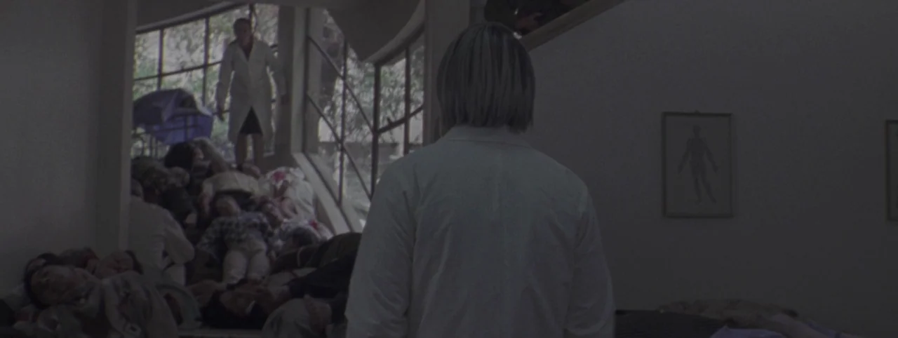 Post Mortem (2010) movie still: medium — A sterile interior hallway features an anatomical diagram on the plain off-white wall. Multiple individuals…