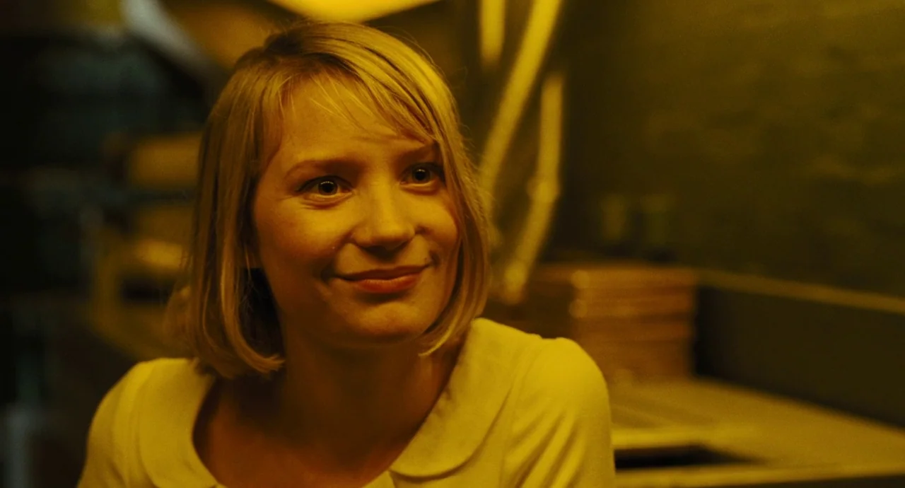 The Double (2013) movie still: closeup — An interior space is illuminated by a yellow light source. A young woman with a blonde bob hair cut is…