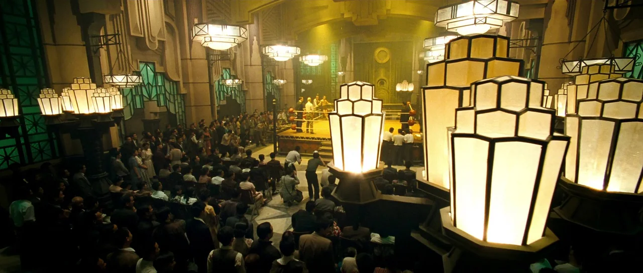 Ip Man 2 (2010) movie still: wide — An opulent 1940s arena with geometric architecture and large ornate hanging light fixtures. A crowd is seated in…