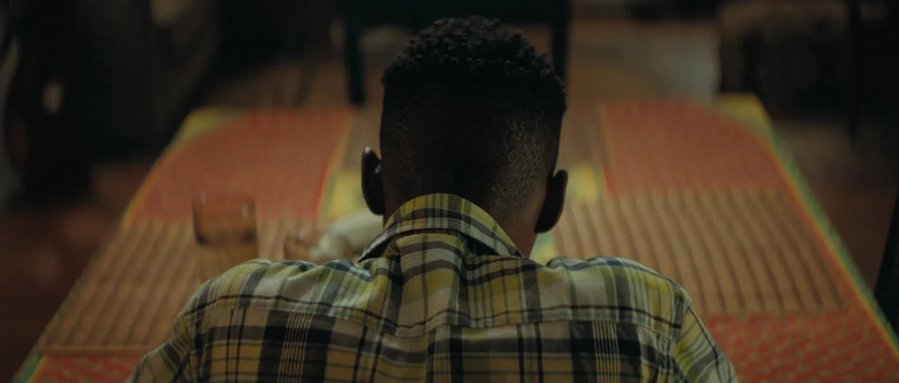 Moonlight (2016) movie still: medium — A man sits at a table covered in a patterned yellow and red cloth. The person faces away from the camera in a…
