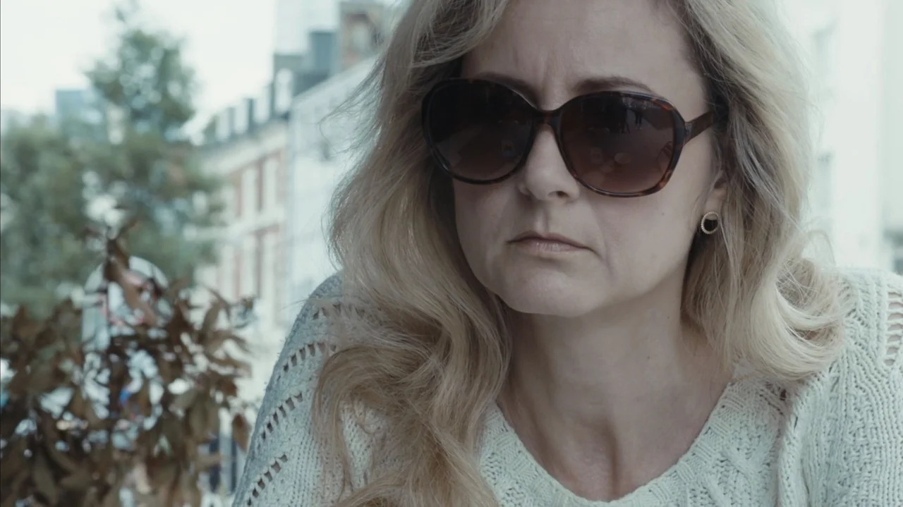 You Might Get Lost (2021) movie still: closeup — An outdoor cafe area with blurred urban architecture visible in the background. A blonde woman in a…