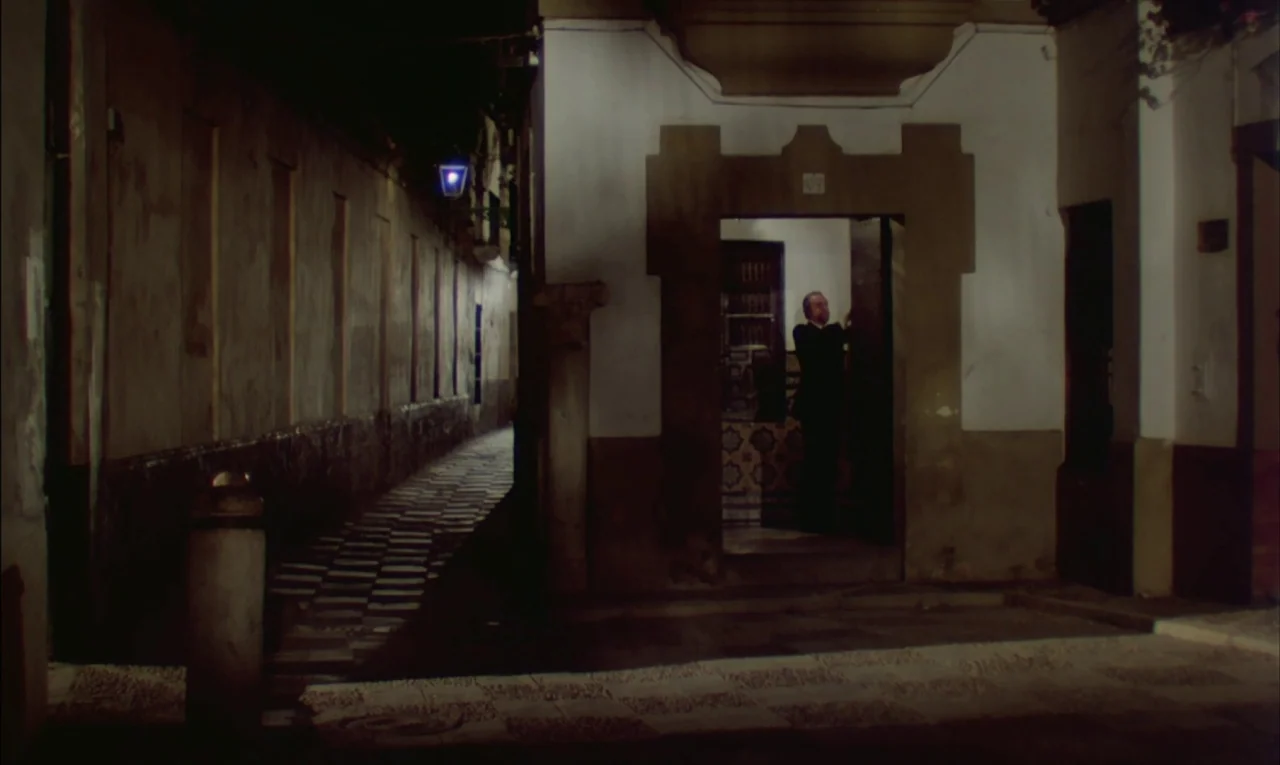 That Obscure Object of Desire (1977) movie still: wide — A narrow stone alleyway at night featuring a white building with an open door. An older man…