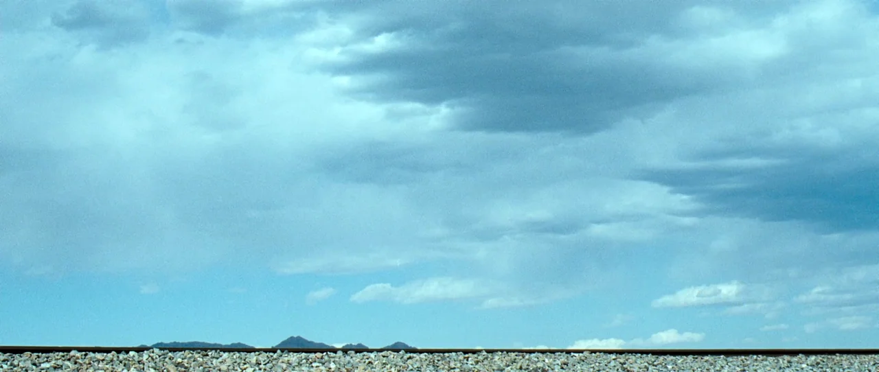 Little Birds (2011) movie still: establishing — An expanse of grey gravel bed borders train tracks under a bright blue sky. Low mountains are…