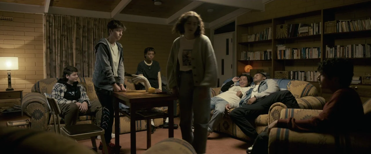 Talk to Me (2023) movie still: wide — A living room featuring paneled walls and bookshelves filled with various books. A group of teenagers…