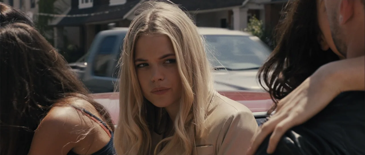Carrie (2013) movie still: medium — A suburban street with a parked silver car and residential houses visible in background. A young woman with long…