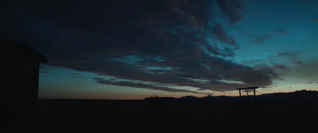 The Wind (2018) movie still: establishing — A vast desert expanse is under a twilight sky with streaky dark blue clouds. A silhouette of a small…