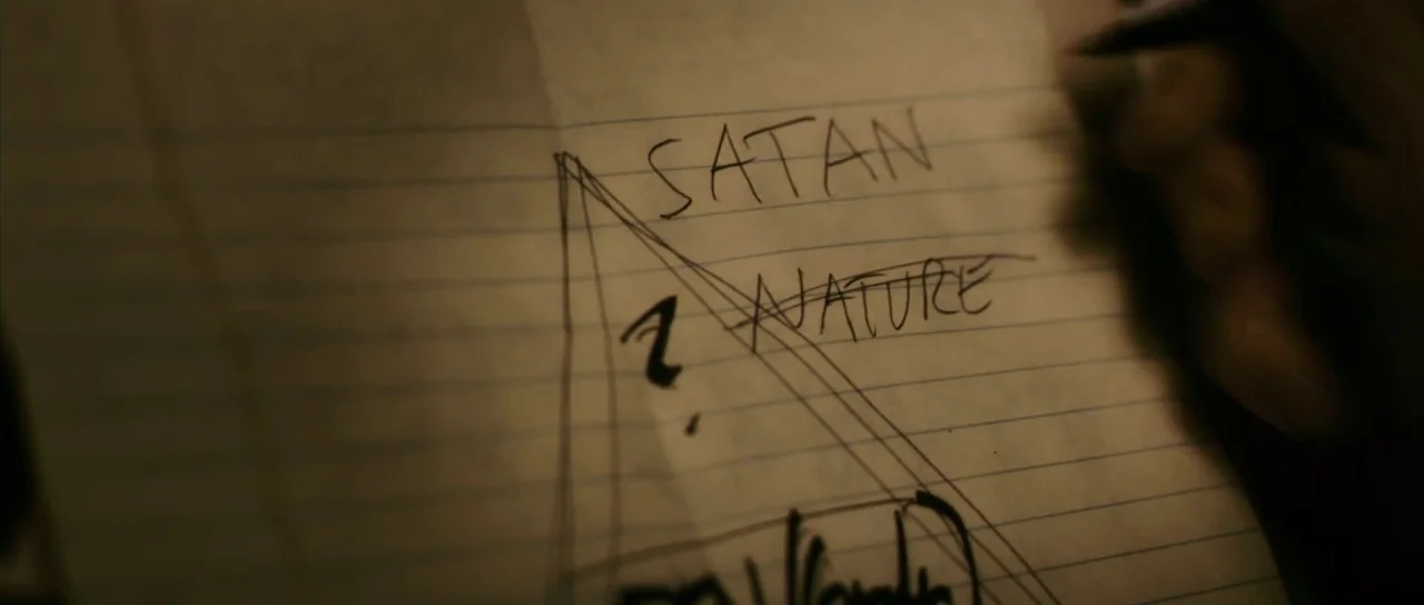 Antichrist (2009) movie still: closeup — A sheet of lined notebook paper rests on a dark surface in dim light. The words Satan and Nature are…