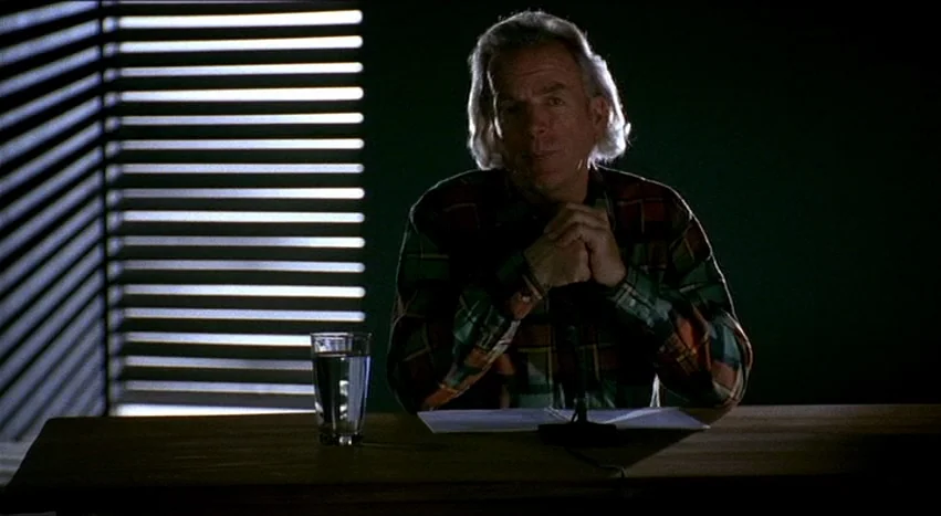 Gray’s Anatomy (1996) movie still: medium — A dimly lit room features a subject sitting behind a wooden table. A glass of water sits next to a…