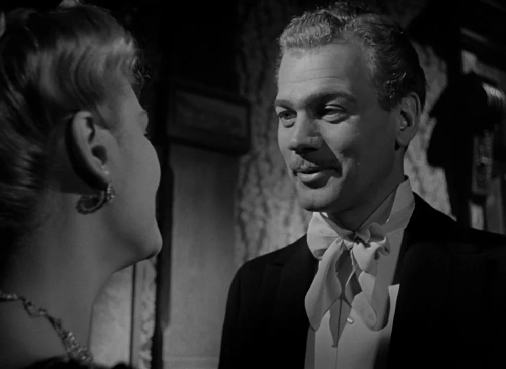 The Magnificent Ambersons (1942) movie still: medium — A dimly lit room with ornate wallpaper serves as the background for the two figures. A man in…
