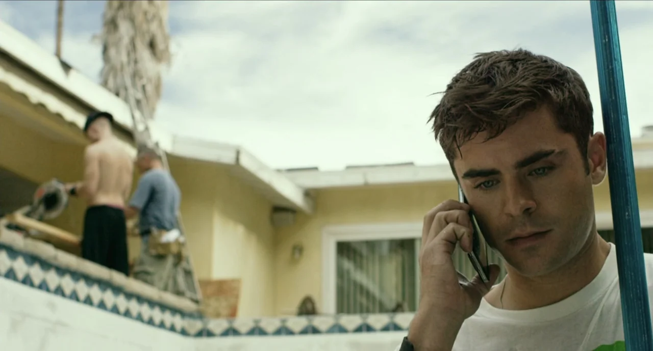 We Are Your Friends (2015) movie still: medium — A sunny outdoor patio area features a yellow stucco wall and tiled border. A young man in a white…