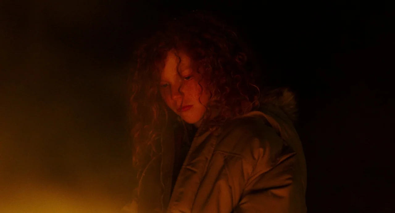 My Animal (2023) movie still: medium — A person stands in total darkness at night. The subject's face and jacket are illuminated by a warm fire.