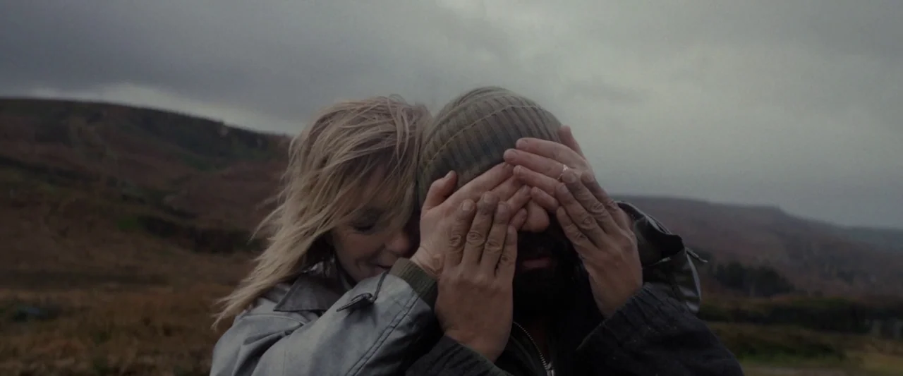 Ali & Ava (2021) movie still: medium — A desolate hillside landscape under a grey, overcast sky with dry brown vegetation. A woman stands behind a…