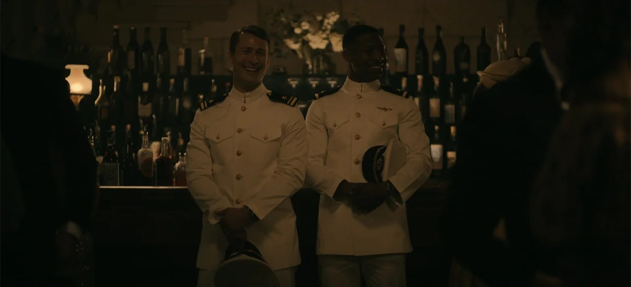 Devotion (2022) movie still: medium — A dimly lit indoor bar features shelves of bottles lined up against the wall. Two naval officers in white…