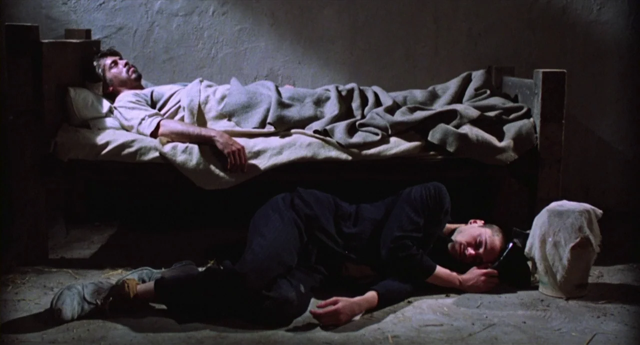 Caravaggio (1986) movie still: medium — A dark, sparse room with textured stone walls and a single wooden bed frame. One man rests on the bed frame…