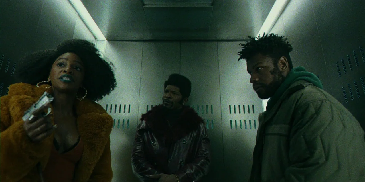 They Cloned Tyrone (2023) movie still: medium — Three individuals stand close together inside a sterile, metallic, cramped box. The woman holds a…