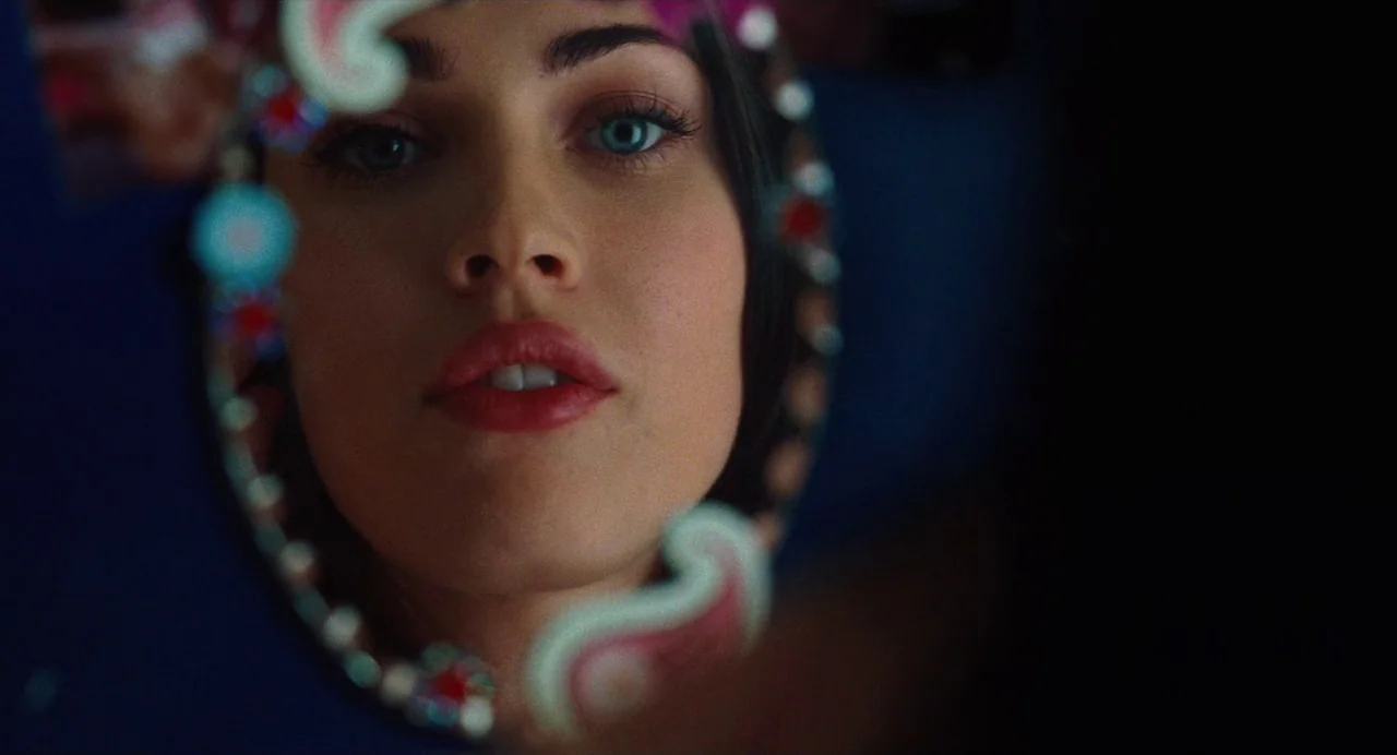 Jennifer’s Body (2009) movie still: closeup — A dark, deep blue environment surrounds a reflective surface containing an illuminated face. A young…