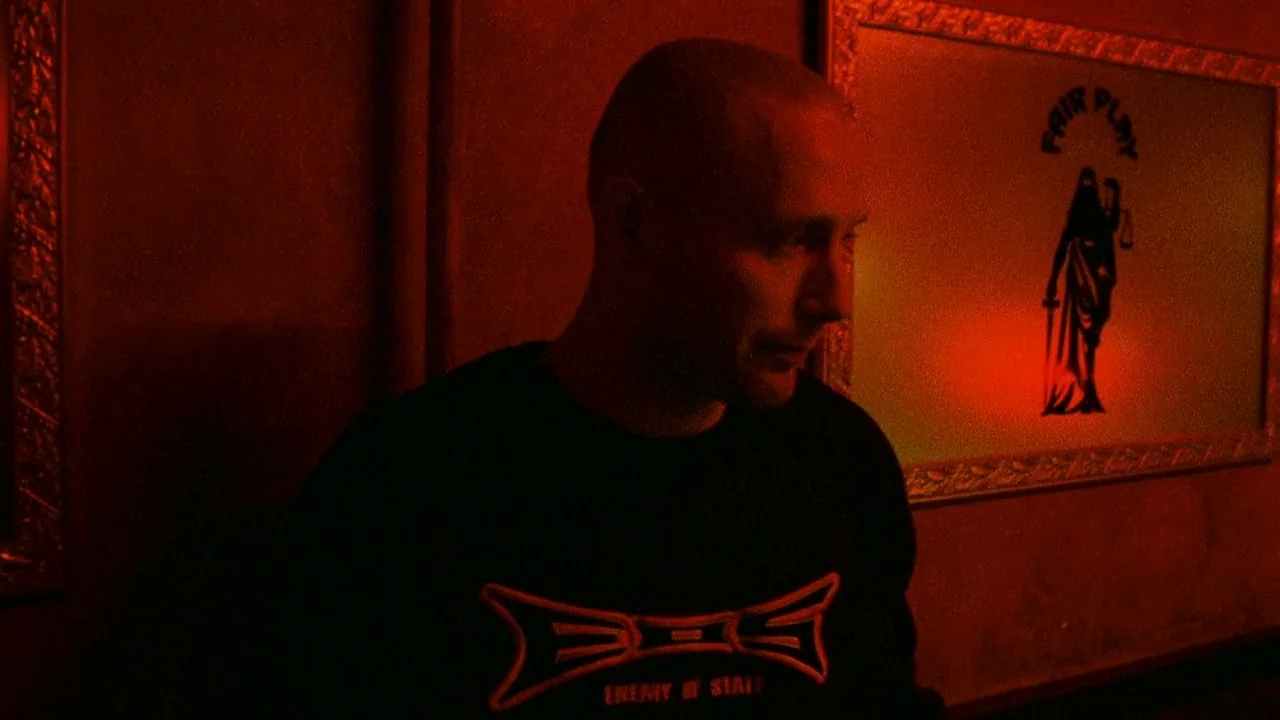 Pusher II (2004) movie still: medium — A dimly lit room with walls washed in deep red light features a framed art piece. A bald man wearing a black…