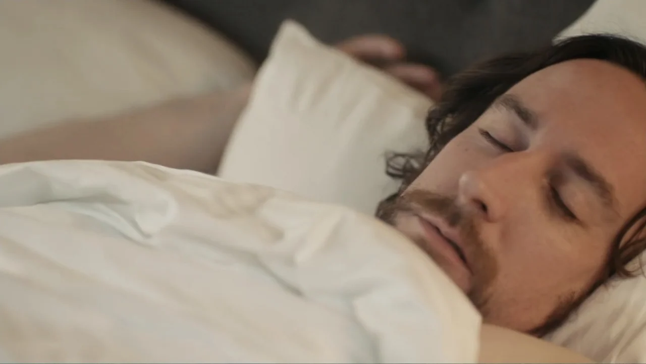 Toss It (2019) movie still: closeup — A man lies in a bed with white sheets and pillows.