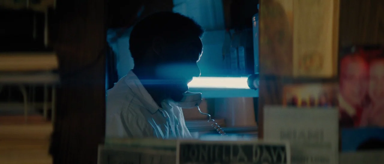 Moonlight (2016) movie still: medium — A dimly lit retail space features a man working behind a cluttered counter area. He holds a white landline…