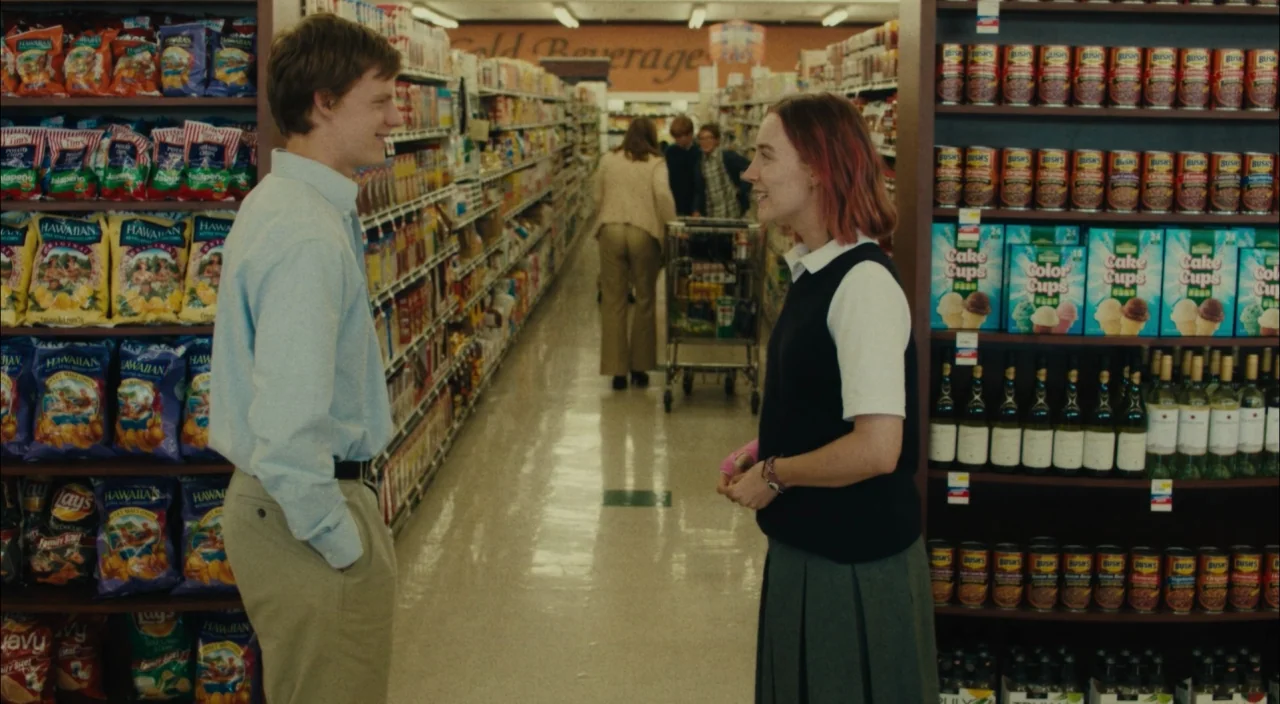 Lady Bird (2017) movie still: medium — An aisle in a grocery store filled with shelves of snack bags and canned goods. A young man and a woman face…