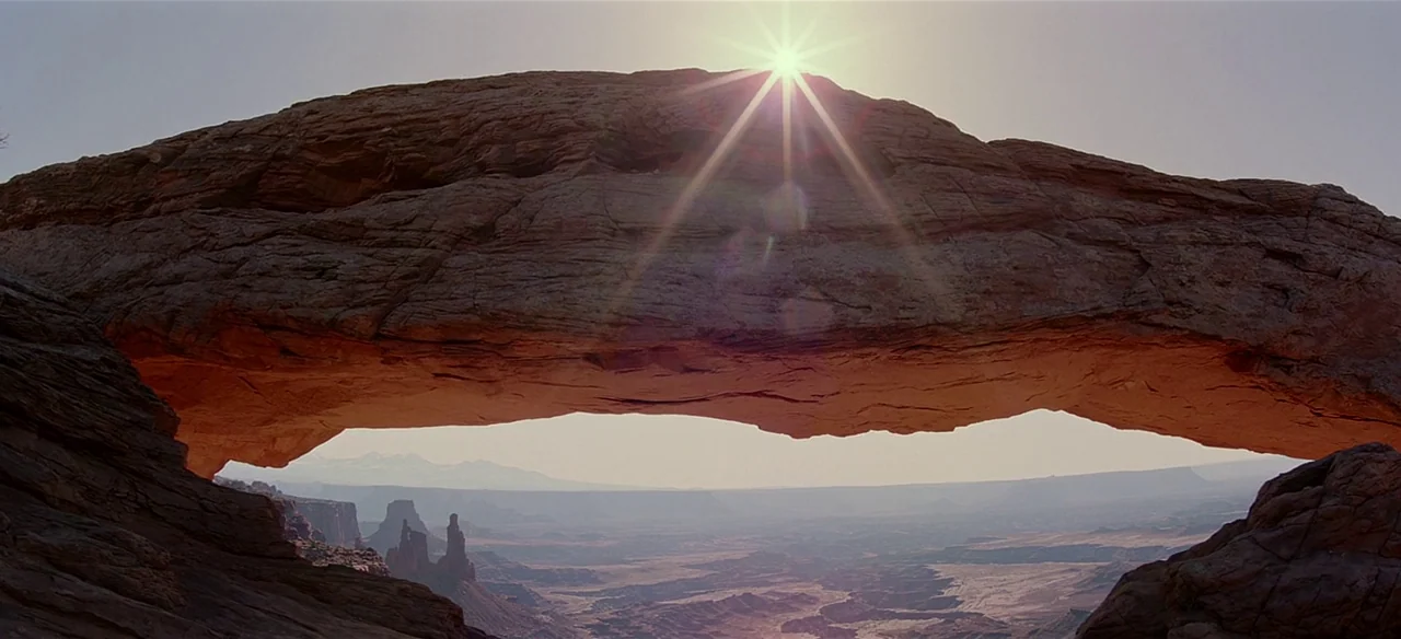 Baraka (1992) movie still: establishing — A massive natural sandstone arch frames an expansive arid desert valley under bright sun. The sun is…