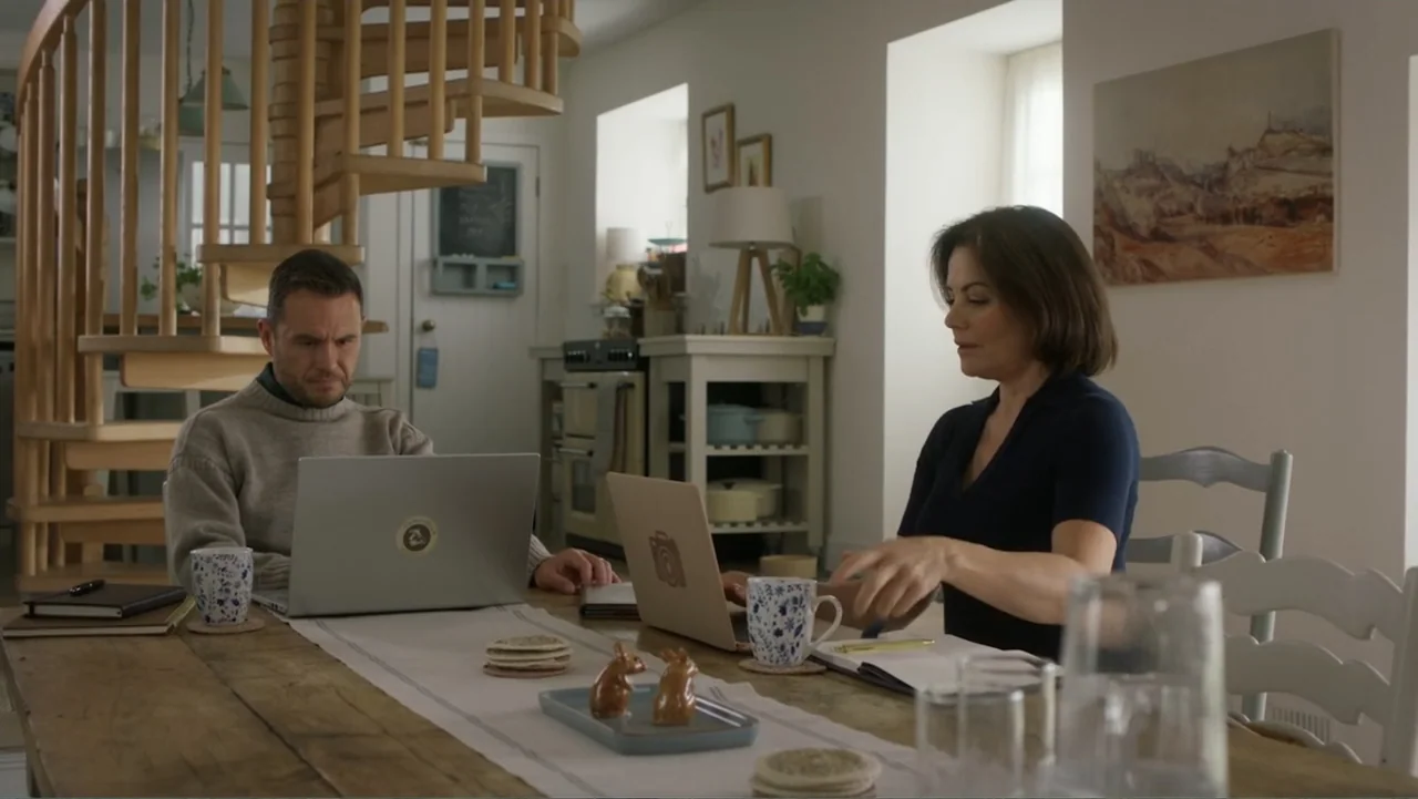 A Scottish Love Scheme (2024) movie still: medium — A wooden dining table sits before a white wall with a spiral staircase background. A man and a…