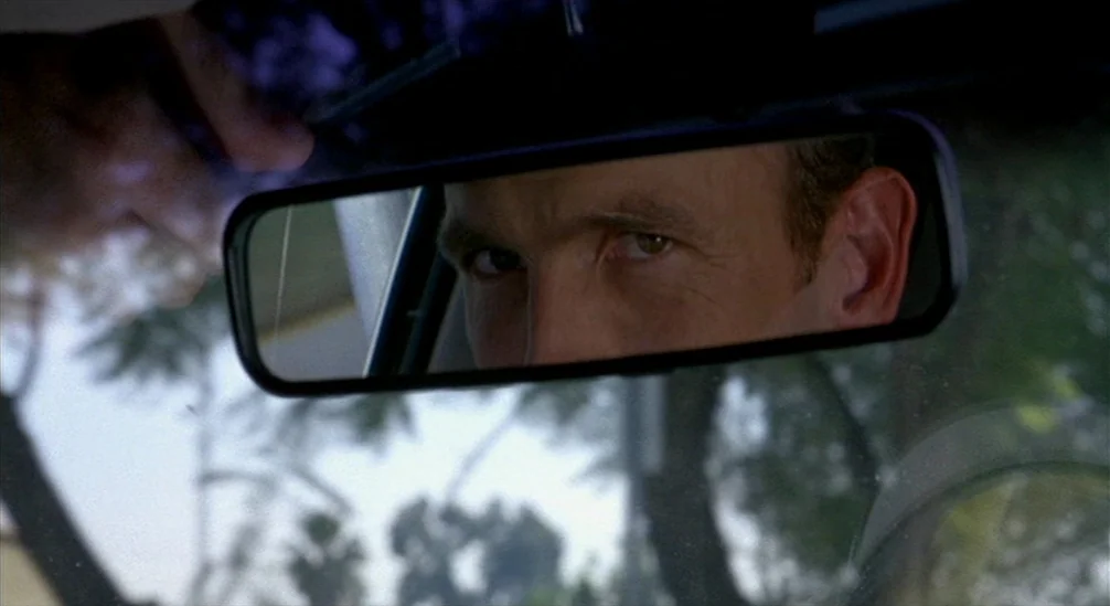 New Nightmare (1994) movie still: closeup — A man's eyes are visible in the reflection of a car rearview mirror. The blurred view through the…