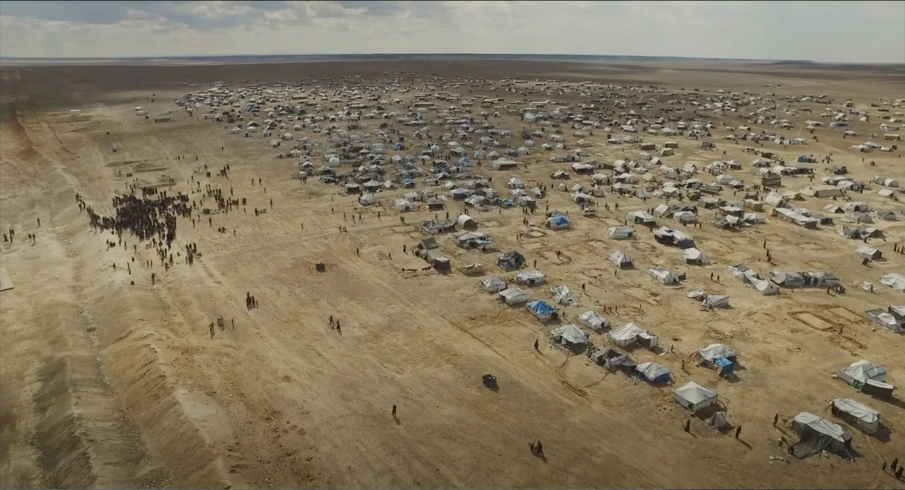 Human Flow (2017) movie still: establishing — A vast, expansive refugee camp covers a flat, dry desert plateau under a cloudy sky. Numerous small…
