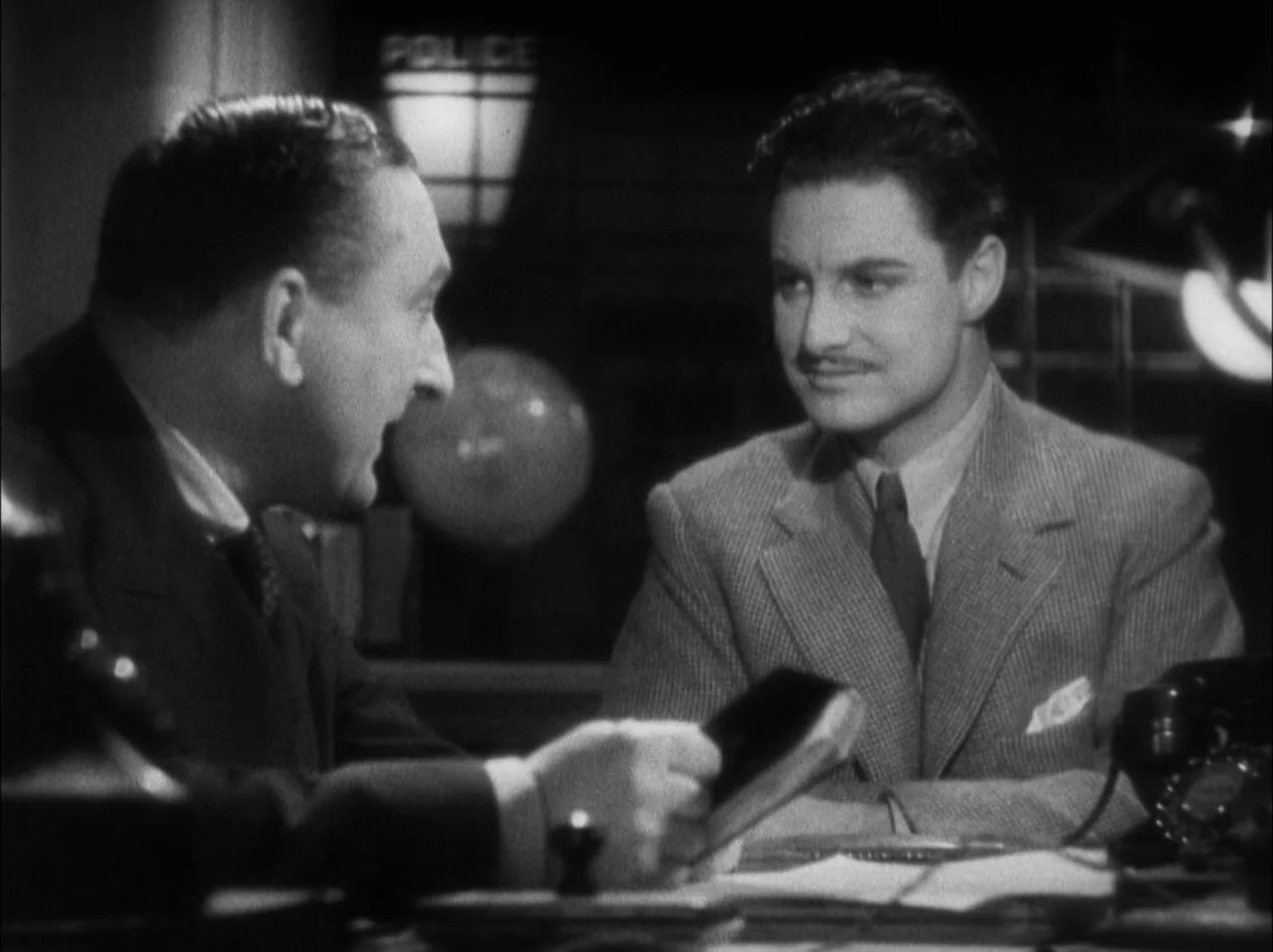 The 39 Steps (1935) movie still: medium — A dimly lit room features a desk with a telephone and a book. Two men in formal business attire face each…