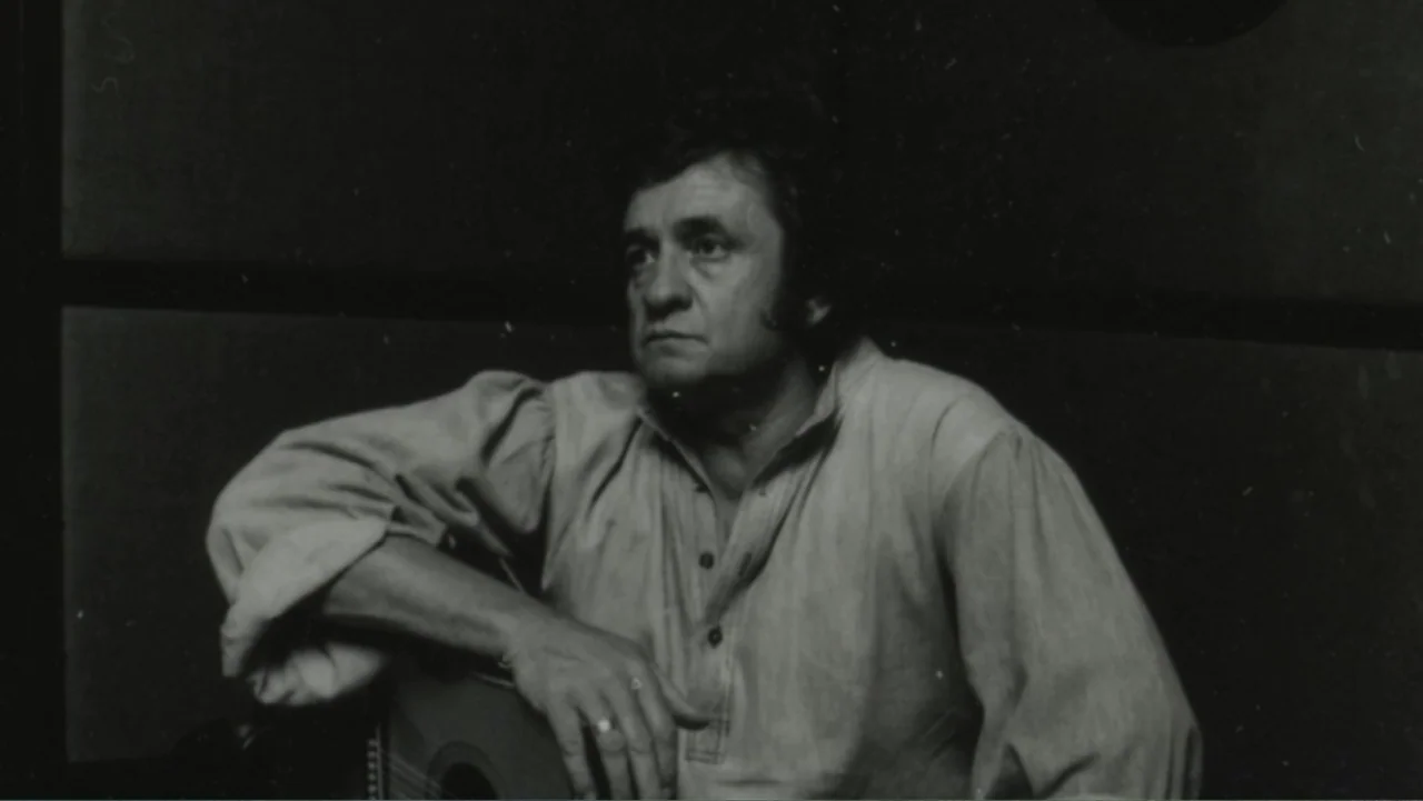 The Winding Stream (2014) movie still: medium — A dimly lit room with plain walls serving as a backdrop. A man holding an acoustic guitar sits while…
