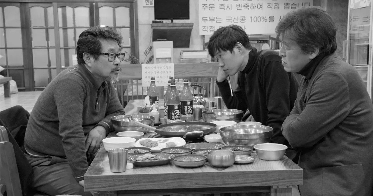 Hotel By The River movie still: medium — A restaurant interior with a wooden table filled with stainless steel bowls and bottles. Three Asian men…