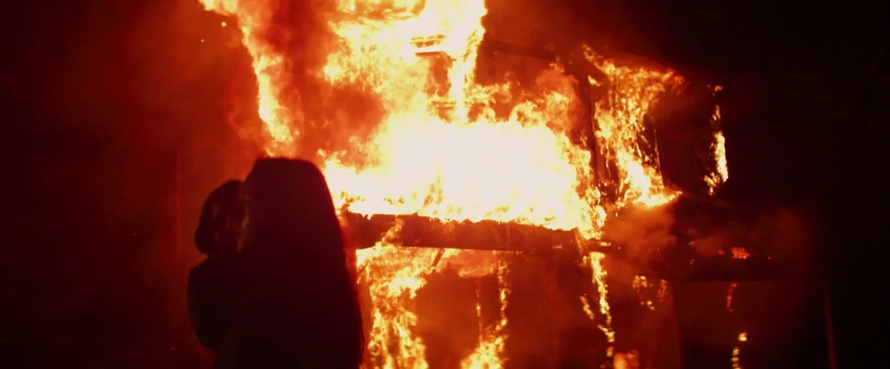 Lost River (2014) movie still: medium — A dark night exterior features a multi-story building engulfed in bright, roaring flames. Two silhouettes…