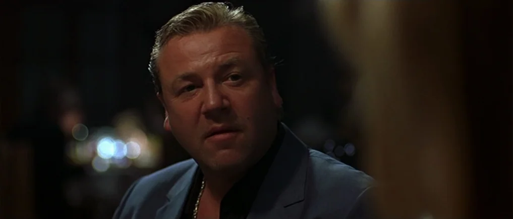 Sexy Beast (2000) movie still: closeup — A man sits in a dimly lit space with blurry bokeh lights in the background. The man looks toward the side…