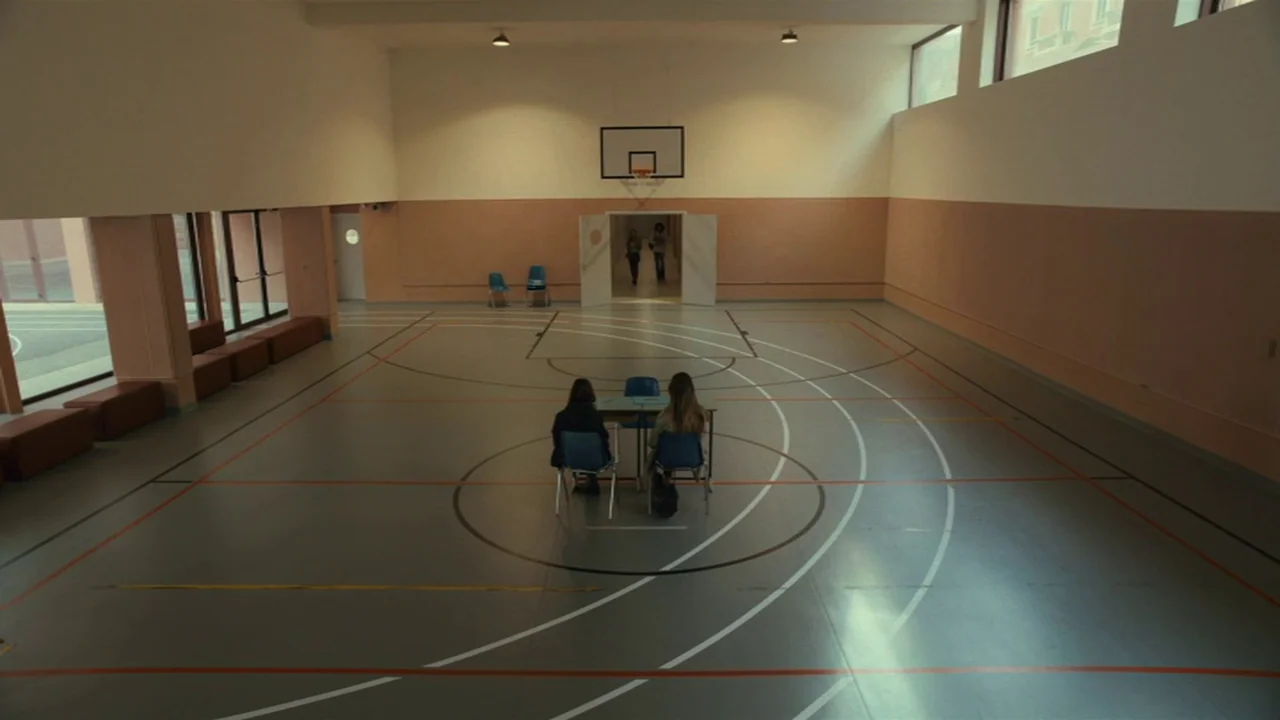 Amanda (2022) movie still: wide — A spacious gymnasium features pale walls and floor markings for basketball and track events. Two students sit at a…