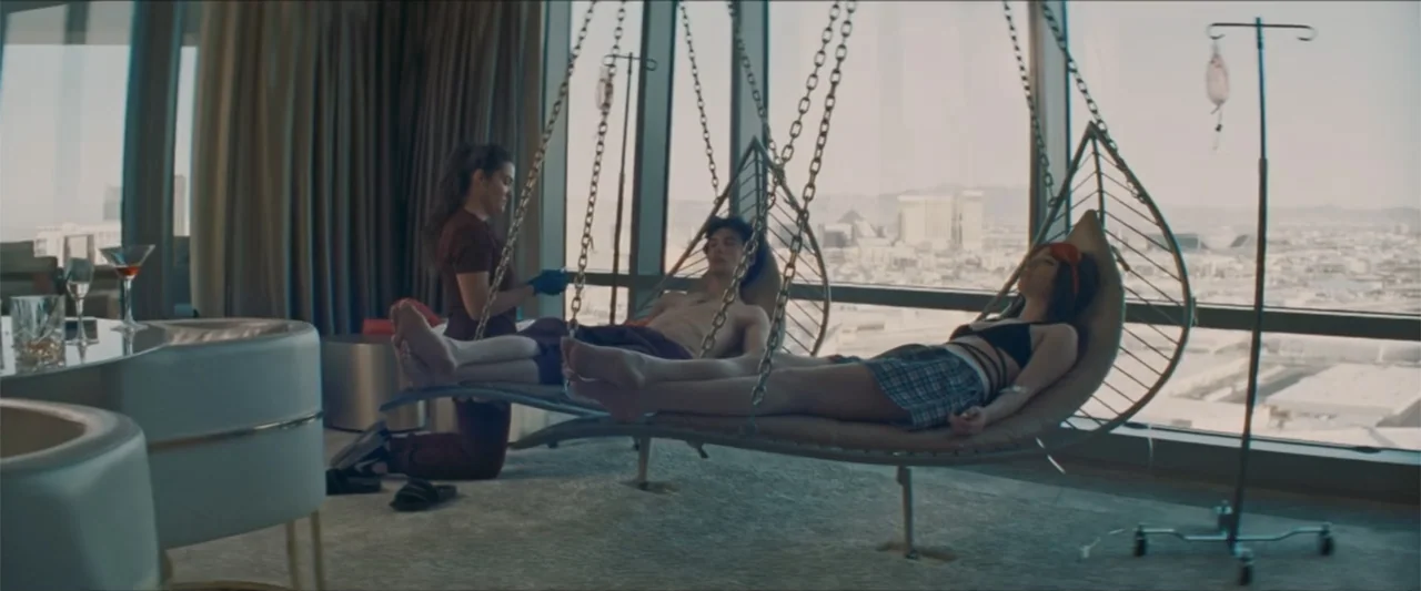 Anora (2024) movie still: wide — A luxury hotel room with panoramic city views and floor-to-ceiling windows. A medical professional provides care to…
