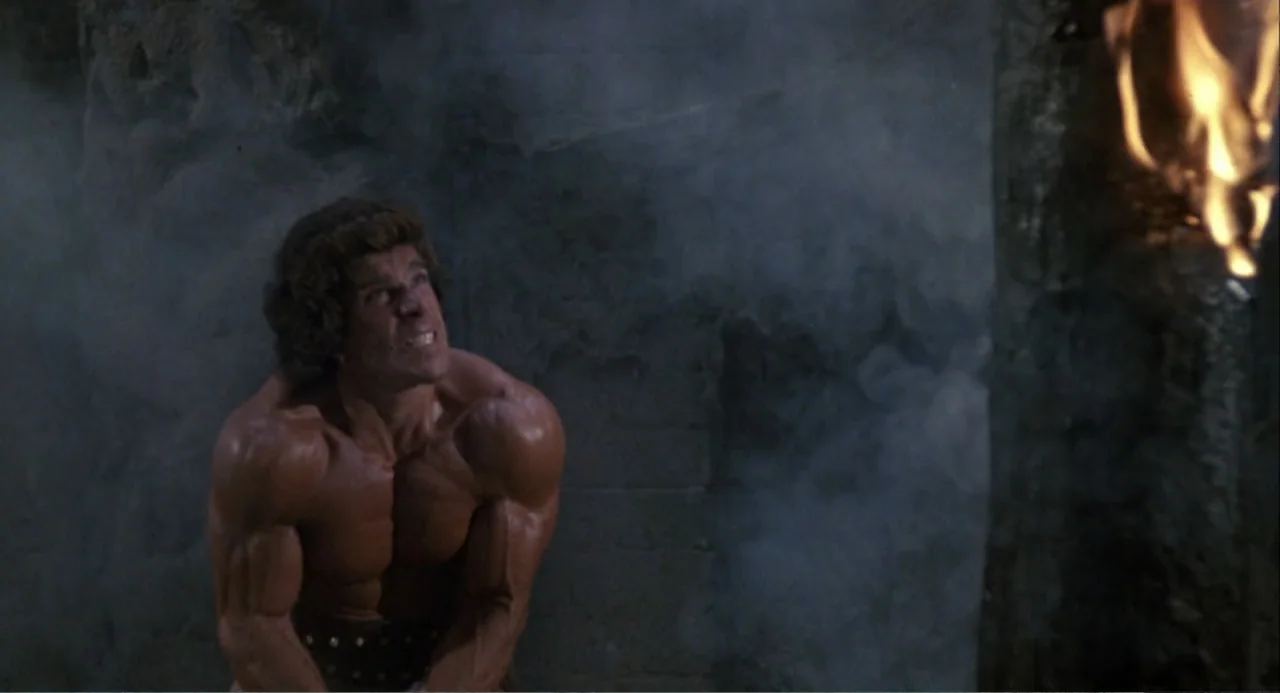 Hercules (1983) movie still: medium — A stone dungeon wall is obscured by thick gray smoke and dim shadows. A muscular man with a strained…