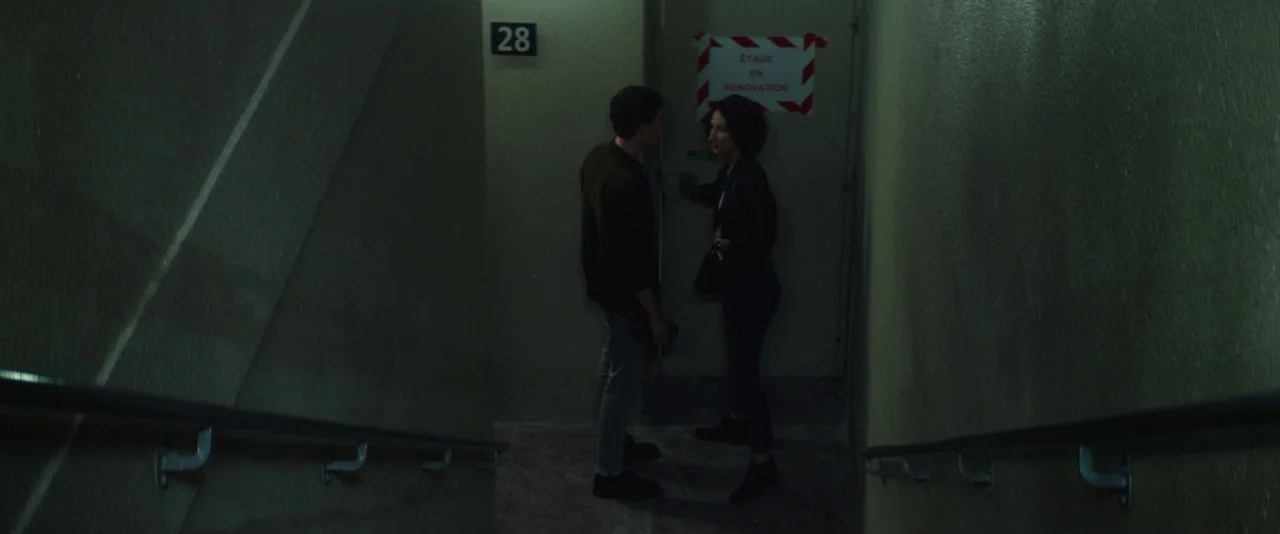 Nocturama (2016) movie still: fullbody — A dimly lit stairwell landing features a door labeled with a red renovation sign. A man and woman stand…