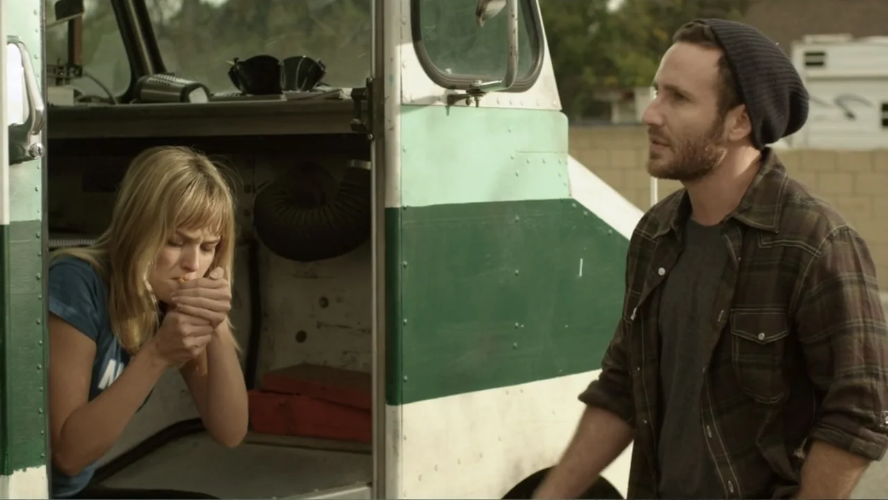 Free Samples (2013) movie still: medium — A woman sits in the open cab of a painted industrial truck. A man stands beside the door looking toward…