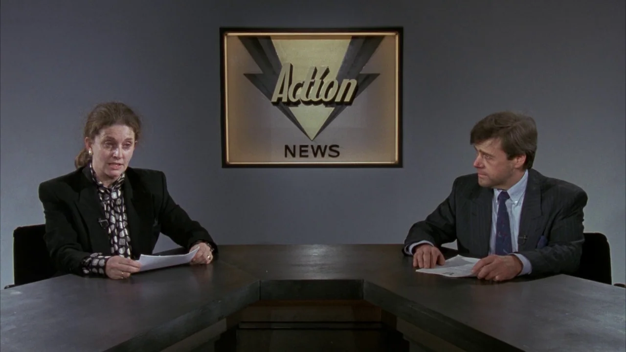 Batman (1989) movie still: medium — A television news studio features a dark grey desk and matching wall panels. A male and a female news anchor sit…