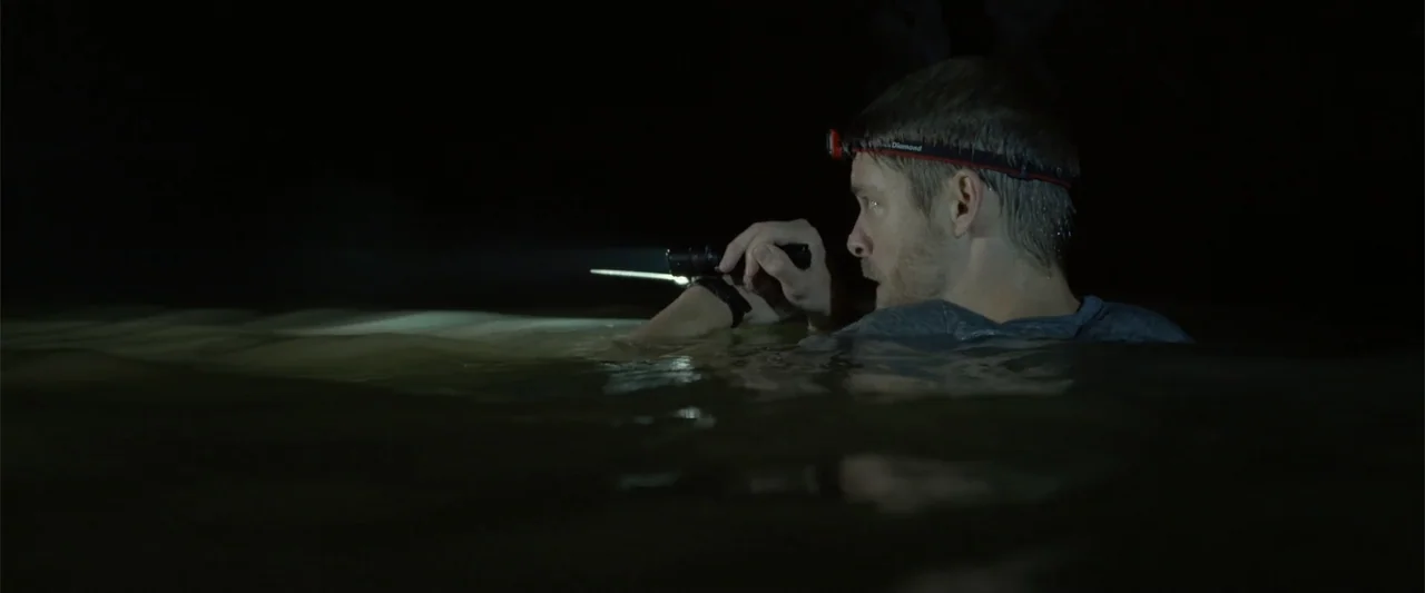 Black Water: Abyss (2020) movie still: medium — A man wades through dark, murky water in a pitch-black outdoor environment. He wears a headlamp and…