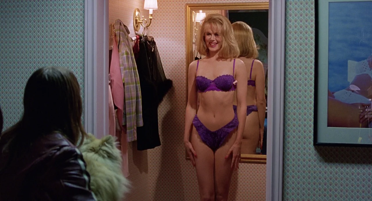 To Die For (1995) movie still: medium — A dressing room features patterned wallpaper and a gold-framed wall mirror. A woman in purple lingerie looks…