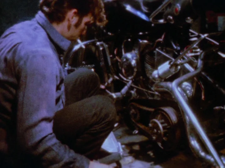 Scorpio Rising (1963) movie still: medium — A dimly lit industrial space containing a man working on a disassembled motorcycle. The man crouches on…