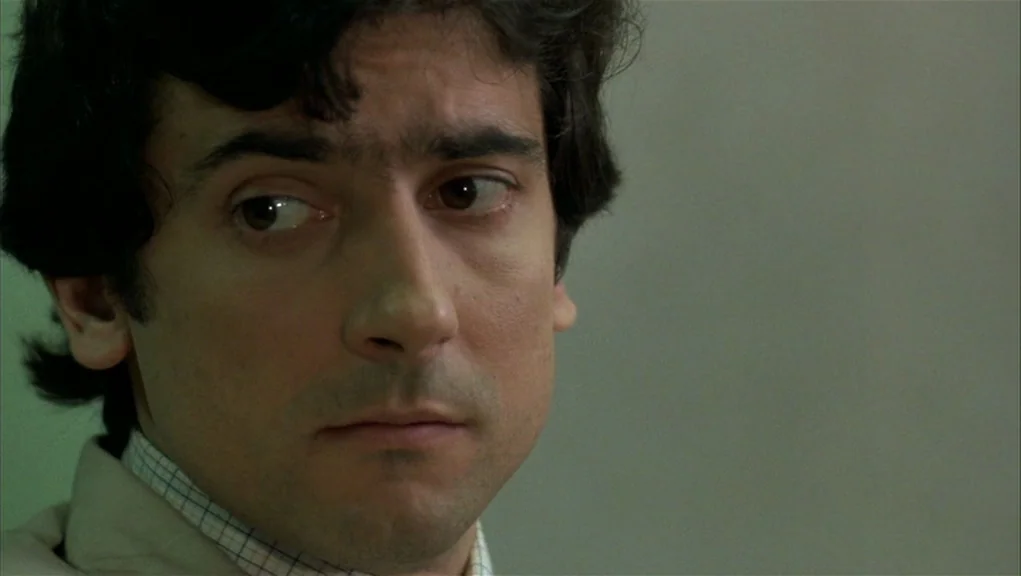 After Hours (1985) movie still: closeup — A man stands in an interior room with a plain, light-toned wall. The man stares intently towards the left…