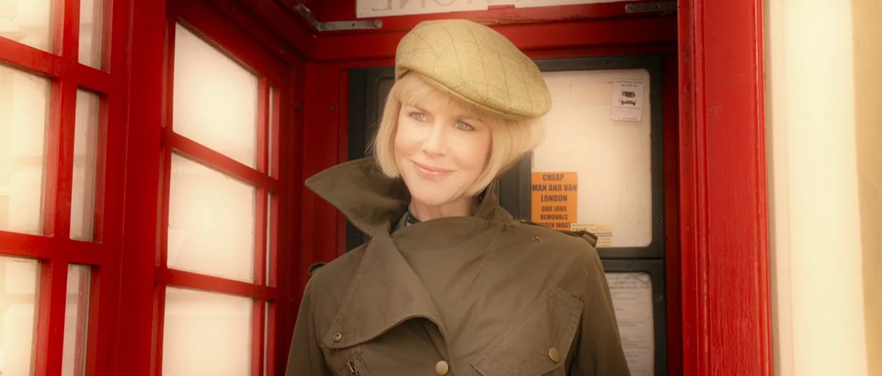 Paddington (2014) movie still: medium — A woman stands inside a bright red glass telephone box. She wears a trench coat and cap while looking out.